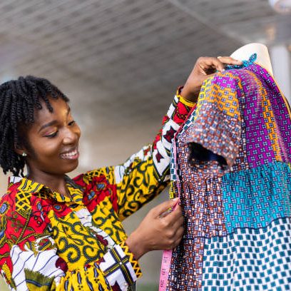 Smiling Ghanaian Dressmaker with locs hair measures a part of her colourful African pattern dress in her studio